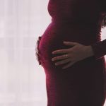 Schwangere Frau in einem roten Kleid die sich mit beiden Händen sanft den Bauch hält