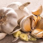 3 Knoblauchknollen. Eine davon ist aufgebrochen wodurch einzelne Zehen zu sehen sind. Eine Zehe ist aufgeschnitten.